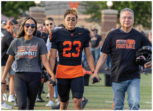 Two people are holding hands, one wearing a black and orange football uniform with the number 23 and the other wearing a black shirt with “West De Pere Phantoms Football" printed on it. Description generated by AI