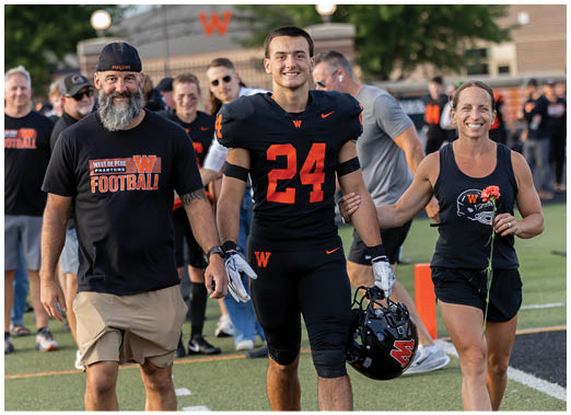 An adult wearing a black sports uniform with the number 24 is walking arm-in-arm with another adult in black athletic clothing holding a flower on a sports field. Description generated by AI
