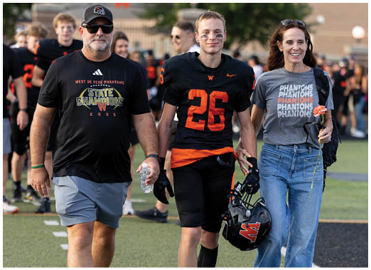 A person wearing a black and orange sports uniform with the number 26 walks beside another person in a gray shirt with repeated “PHANTOMS" text and blue jeans holding a flower. Description generated by AI