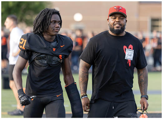A person wearing a black t-shirt with a red heart design and a white patch that says “CREATE" stands next to another person in a black sports uniform on a field. Description generated by AI