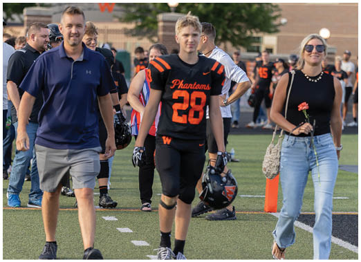 An adult in a black and orange football uniform walks on a field holding a helmet while another adult in casual clothing holds a flower and a woven bag. Description generated by AI