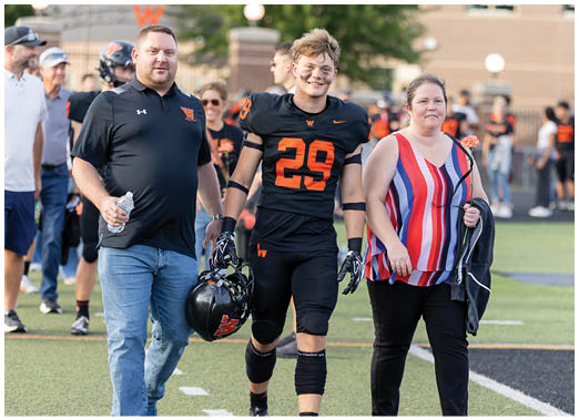 An adult wearing a striped sleeveless top and holding a flower and jacket walks beside a person in a black and orange football uniform on a sports field. Description generated by AI