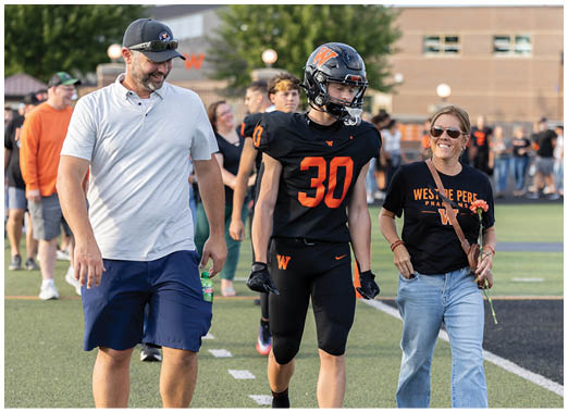 An adult wearing sunglasses and a black Westside Peregrine t-shirt walks on a sports field holding a flower next to a person in a black football uniform with the number 30. Description generated by AI