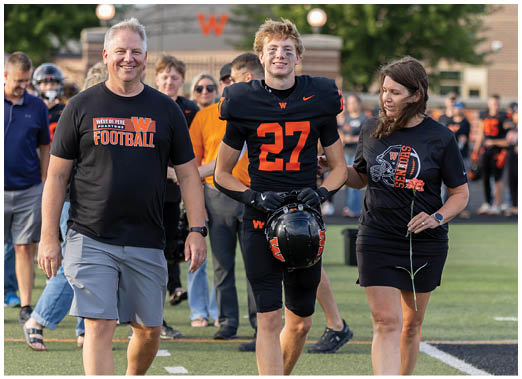 An adult in a black football uniform holding a helmet stands next to another adult wearing a black shirt with a football graphic and the word “Seniors" while holding a flower on a sports field. Description generated by AI