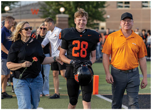 A person in a black football uniform with the number 28 is holding a helmet and standing next to a person wearing an orange polo shirt on a sports field. Description generated by AI