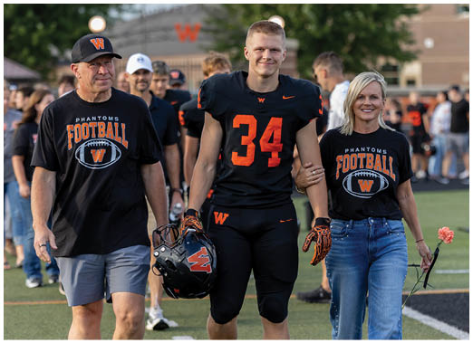 An adult wearing a black football uniform with the number 34 is walking arm-in-arm with another adult wearing a black “Phantoms Football" t-shirt and jeans while holding a flower. Description generated by AI