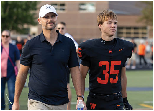 A person wearing a black sports jersey with the number 35 stands next to another person in a dark blue polo shirt outdoors. Description generated by AI
