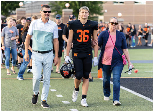 An adult wearing a black sports uniform with the number 36 is walking arm-in-arm with another adult dressed in casual clothing holding a water bottle and sunglasses on a sports field. Description generated by AI