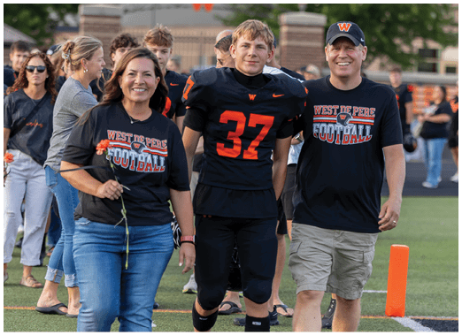 Two people are walking side by side on a sports field, one wearing a black football uniform with the number 37 and the other wearing a black West De Pere Football t-shirt and light-colored shorts. Description generated by AI