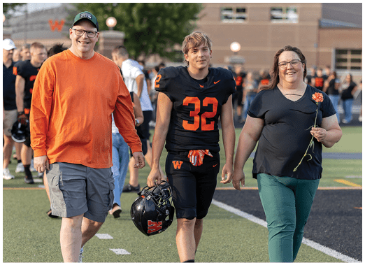 An adult wearing a black sports jersey with the number 32 walks beside another adult holding a flower on a sports field with blurred people in the background. Description generated by AI