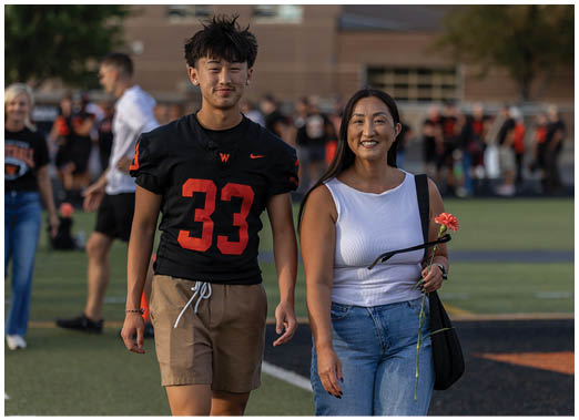 A person wearing a white sleeveless top and jeans is smiling while holding a flower and standing next to another person in a sports uniform on a field with blurred spectators in the background. Description generated by AI