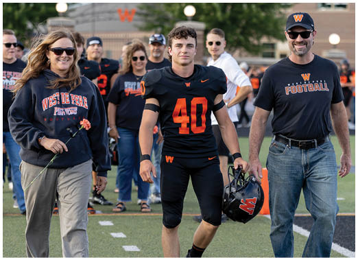 An adult wearing a black football uniform with the number 40 stands next to another adult in a black West Perry Football t-shirt and jeans, holding a football helmet on a sports field. Description generated by AI