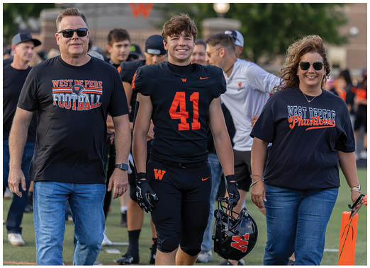 A person wearing a black sports uniform with the number 41 stands next to a person in a navy blue shirt with “West De Pere Phantoms" written on it, both outdoors. Description generated by AI