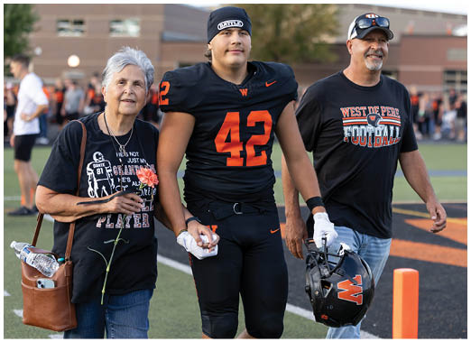 Two people stand side by side on a sports field, one wearing a black and orange football uniform with the number 42 and the other wearing a black West De Pere Football t-shirt and holding a helmet. Description generated by AI