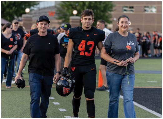An adult wearing a dark sports uniform with the number 43 stands next to another adult in casual clothing holding a single red flower on a sports field. Description generated by AI