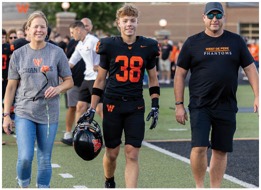 A person wearing a black and orange football uniform with the number 38 stands next to another person in a black West De Pere Phantoms t-shirt on a sports field. Description generated by AI