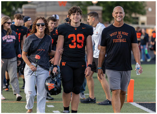 Two people walk side by side on a sports field, one wearing a black football uniform with the number 39 and the other wearing a black West De Pere Football t-shirt and gray pants. Description generated by AI