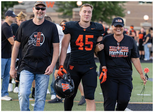 A person wearing a football uniform with the number 45 is walking arm-in-arm with another person wearing a West De Pere Football t-shirt and holding a flower. Description generated by AI