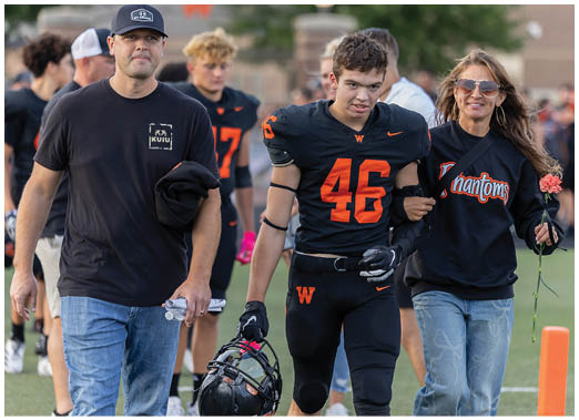 A person wearing a black football uniform with the number 46 in orange is being supported by another person wearing a black sweatshirt with the word “Phantoms" on it. Description generated by AI