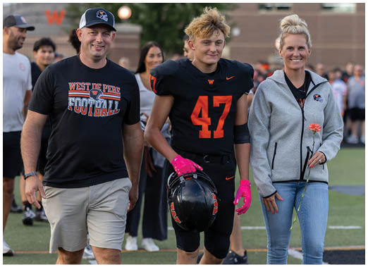 A person wearing a black football uniform with the number 47 and pink gloves is holding a black helmet while standing next to another person in a gray zip-up jacket holding a single orange flower. Description generated by AI