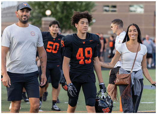 An adult wearing a black football uniform with the number 49 is being accompanied by another adult in a white shirt and gray sweater tied around the waist, smiling and holding the player's arm on a sports field. Description generated by AI