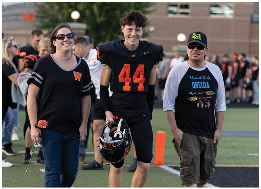 Two people stand side by side on a sports field, one wearing a black football uniform with the number 44 and holding a helmet, and the other wearing sunglasses, a cap, and a black and white shirt with colorful text and designs. Description generated by AI