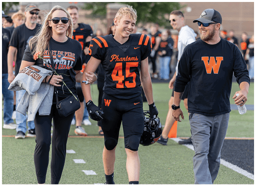 An adult wearing a black football uniform with the number 45 and the word Phantoms walks next to another adult in a black long-sleeve shirt with an orange W, holding a water bottle on a sports field. Description generated by AI
