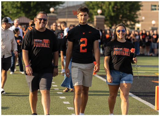 Two people walk side by side on a sports field, one wearing a black football jersey with the number two and the other wearing a black football-themed t-shirt and holding an orange flower. Description generated by AI