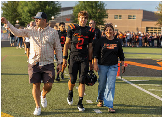 An adult wearing a West De Pere Football sweatshirt walks beside a person in a black and orange Phantoms football uniform holding a helmet on a sports field with a crowd in the background. Description generated by AI