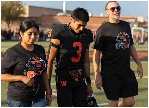 Two people stand on a sports field wearing black athletic clothing, one in a football uniform with the number three and gloves, and the other in a black shirt with a sports logo. Description generated by AI