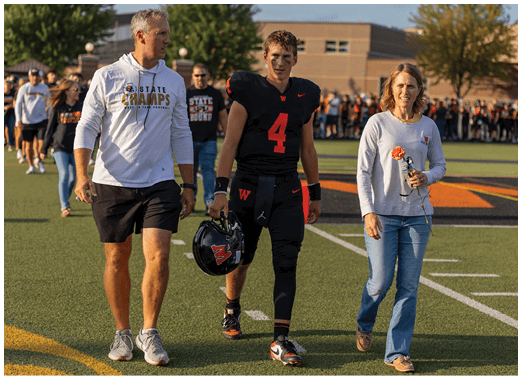 A person in a black sports uniform holding a helmet walks beside a person in casual clothing holding a flower and a water bottle on a sports field. Description generated by AI