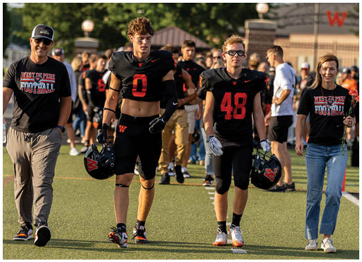 People wearing black football uniforms with red numbers are walking on a field with others in casual clothing in the background. Description generated by AI