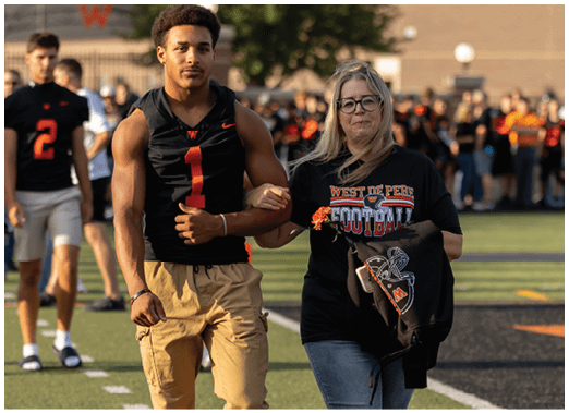 A person wearing glasses and a West De Pere Football shirt holds a flower and links arms with another person on a sports field with a crowd in the background. Description generated by AI