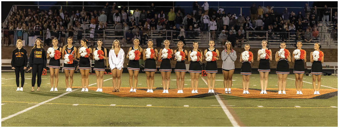 A group of people in cheerleading uniforms and casual clothing stand in a line on a football field near the 50-yard line with a crowd in the background. Description generated by AI