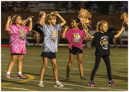 Three children wearing West De Pere Dance Experience shirts and holding pom-poms perform a cheer routine on a sports field at night. Description generated by AI