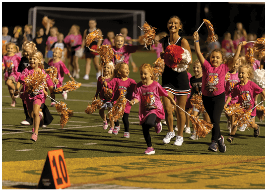 Children wearing matching pink shirts and holding orange pom-poms are running excitedly on a sports field alongside adults in cheerleading outfits. Description generated by AI