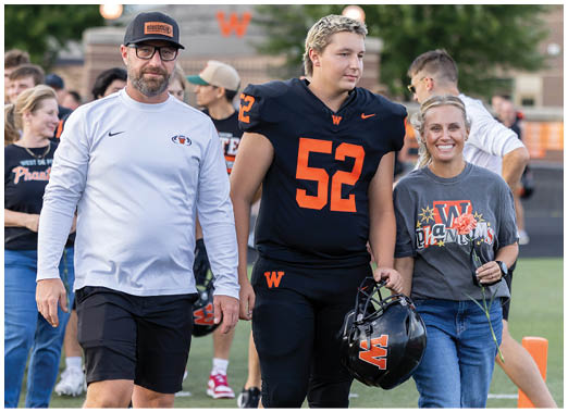 An adult in a football uniform holding a helmet walks on a field between two adults wearing casual clothing, all smiling and surrounded by other people in the background. Description generated by AI