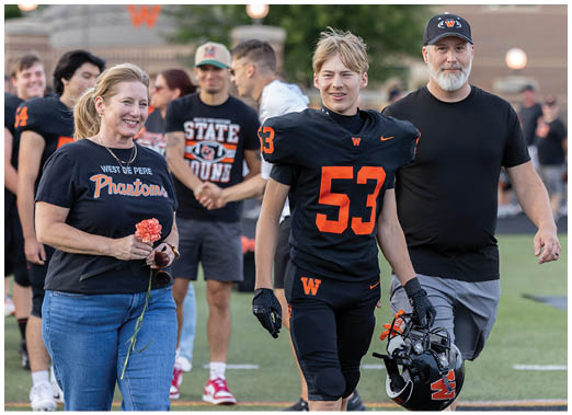 An adult and a person in a football uniform with the number 53 walk together on a sports field while other people stand and interact in the background. Description generated by AI