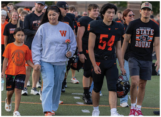 People wearing sports and casual clothing walk together on a field, with one person holding a flower and another holding a football helmet. Description generated by AI