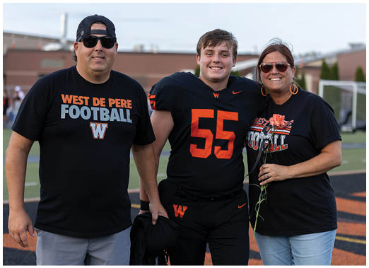 Three people stand together on a football field with one person in a black football uniform and the others wearing black West De Pere Football shirts, with one holding a flower. Description generated by AI