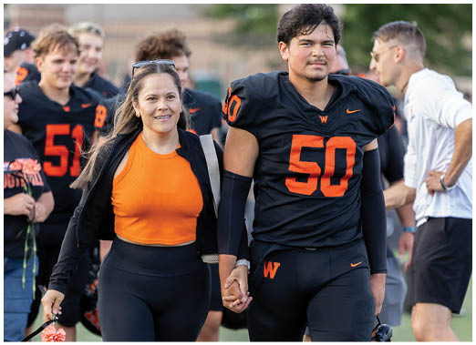 An adult wearing an orange shirt and black jacket walks hand in hand with a person in a black football uniform with the number 50 on it, surrounded by other people in similar sports attire on a field. Description generated by AI