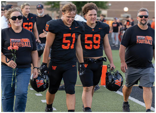 People wearing football uniforms and casual clothing walk together on a sports field, with two holding helmets and one holding flowers. Description generated by AI