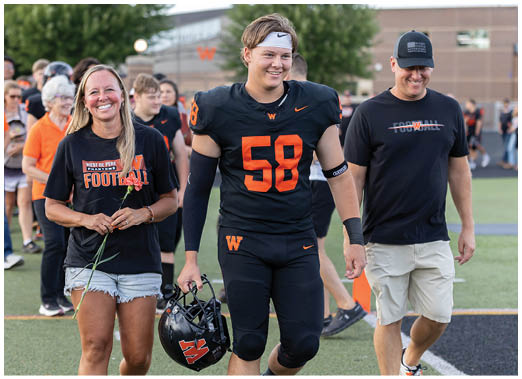 An adult in a football uniform holding a helmet walks arm-in-arm with two adults on a sports field, one holding a flower and wearing sunglasses. Description generated by AI