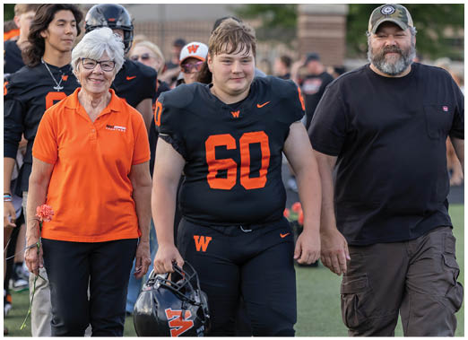 A person in a black and orange football uniform holding a helmet is walking hand in hand with two adults, one wearing an orange polo shirt and the other in a black t-shirt and cap, on a sports field with other people in the background. Description generated by AI
