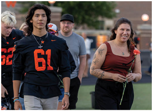 Two adults walk side by side on a sports field, one wearing a black sports jersey with the number 61 and the other holding a flower and smiling. Description generated by AI