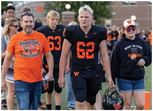 A group of people walk on a sports field with two adults wearing West De Pere Phantoms apparel and two athletes in black football uniforms numbered 62 and 63. Description generated by AI