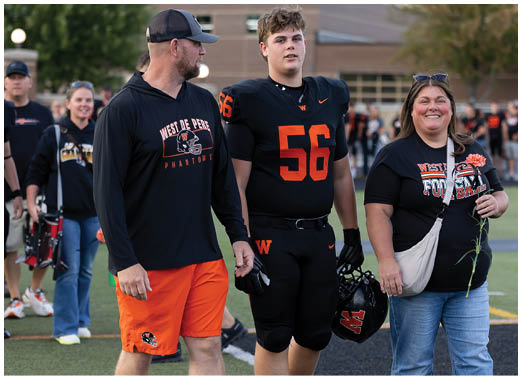 Three people stand on a sports field with one person in a black and orange football uniform holding a helmet, while the others wear casual clothing with team logos and one holds a flower. Description generated by AI