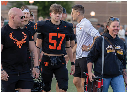 A group of people on a sports field includes a person in a black football uniform with the number 57, another person wearing a black shirt with an orange deer logo, and others dressed casually, some smiling and interacting. Description generated by AI