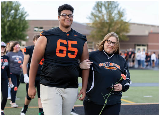 An adult wearing a black sports jersey with the number 65 walks arm-in-arm with another adult holding a flower on a sports field with people and a building in the background. Description generated by AI