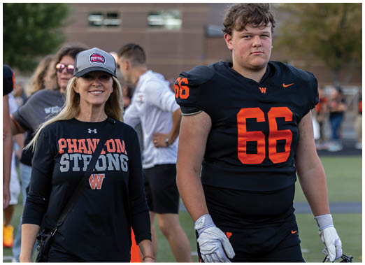 An adult wearing a black football uniform with the number 66 stands next to another adult in a black shirt and cap with “PHANTOM STRONG" printed on it, both on a sports field with other people in the background. Description generated by AI